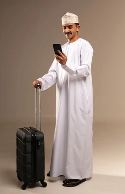 Making a phone call while traveling, looking at something with expressions of excitement and joy, having a good time on vacation, the concept of tourism and travel, a portrait of a young Arab Gulf Omani man wearing a dishdasha and a kumma holding a travel bag in his hand, preparing for travel time, full body, beige background.