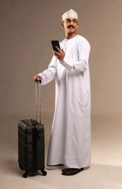 تصفح مواقع التواصل الاجتماعي، النظر الى شيء ما بتعابير الحماس والسرور, قضاء أوقات ممتعة في الإجازة, مفهوم السياحة والسفر، بورتريه لشاب عربي خليجي عماني يرتدي الدشداشة و الكمه يمسك حقيبة السفر بيده, الاستعداد لوقت الرحلة، الجسم بالطول الكامل, خلفية بيج