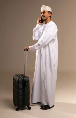 تواصل مع الاهل قبل السفر، النظر الى شيء ما بتعابير الحماس والسرور, قضاء أوقات ممتعة في الإجازة, مفهوم السياحة والسفر، بورتريه لشاب عربي خليجي عماني يرتدي الدشداشة و الكمه يمسك حقيبة السفر بيده, الاستعداد لوقت الرحلة، الجسم بالطول الكامل, خلفية بيج