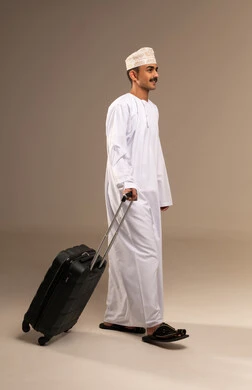 Dragging a suitcase with gestures of happiness and joy, looking at something with expressions of excitement and pleasure, spending enjoyable times on vacation, the concept of tourism and travel, a portrait of a young Arab Gulf Omani man wearing a dishdasha and a kumma holding a suitcase in his hand, preparing for travel time, full-body, beige background.