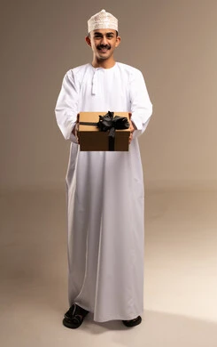 مفهوم الشكر والامتنان، تقديم الهدايا في العيد والمناسبة الخاصة،  التعبير عن الحب والتقدير والاحترام، بورتريه لرجل عربي خليجي عماني يرتدي الدشداشة والكمة يحمل صندوق الهدية ينظر إليه بايماءات المفاجأة والصدمة، الجسم بالطول الكامل، خلفية بيج