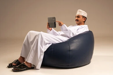 استعمال التكنولوجيا والتقنيات الحديثة، الدردشة مع الأهل والأصدقاء، بورتريه لرجل عربي خليجي عماني يرتدي الدشداشة والكمة يجلس على كرسي مريح يحمل بيده جهاز التابلت، تصفح مواقع التواصل الاجتماعي، استخدام تطبيقات وبرامج الأجهزة اللوحية، خلفية بيج