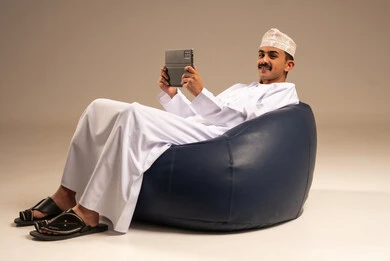 Using tablet applications and programs, utilizing modern technology and techniques, chatting with family and friends, a portrait of an Arab Gulf Omani man wearing a dishdasha and kumma sitting on a comfortable chair holding a tablet device, browsing social media, beige background.