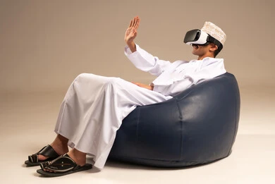 Watching virtual reality movies, immersing and interacting with the imaginary world, using advanced technologies and techniques, a portrait of a young Arab Gulf Omani man wearing a dishdasha and an Omani kumma, wearing VR glasses on his eyes, spending leisure time in an enjoyable way, beige background.