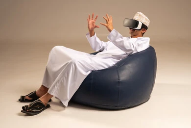 Spending enjoyable and entertaining times with virtual games, enjoying watching virtual reality movies, a portrait from behind of an Arab Gulf Omani man wearing traditional attire sitting on a comfortable chair and wearing 3D virtual reality glasses, gray background.