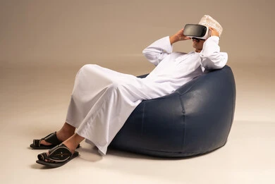 قضاء أوقات ممتعة ومسلية بالألعاب الافتراضية، الاستمتاع بمشاهدة أفلام الواقع الافتراضي، بورتريه من الخلف لرجل عربي خليجي عماني يرتدي الثوب التقليدي يجلس على كرسي مريح ويرتدي نظارة الواقع الافتراضي ثلاثية الابعاد، خلفية رمادية