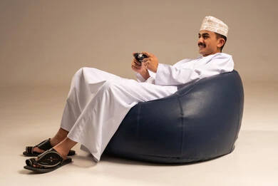 لعب العاب الفيديو، المتعة والتسلية والتفاعل، قضاء وقت ممتع في المنزل، بورتريه لشاب عربي خليجي عماني يرتدي الدشداشة والعمامة  يجلس على كرسي مريح يمسك بيده قبضة التحكم يرفع يده بايماءات النصر، خلفية رمادية