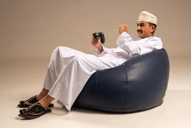 لعب العاب الفيديو، المتعة والتسلية والتفاعل، قضاء وقت ممتع في المنزل، بورتريه لشاب عربي خليجي عماني يرتدي الدشداشة والعمامة  يجلس على كرسي مريح يمسك بيده قبضة التحكم يرفع يده بايماءات النصر، خلفية رمادية