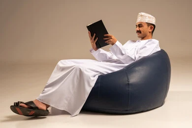 قراءة القصص والروايات, بورتريه لرجل عربي خليجي عماني يرتدي الثوب التقليدي يجلس على كرسي مريح يحمل بيده كتاب يقوم بقراءته, ممارسة هواية المطالعة, الاستمتاع بالأنشطة الترفيهية المفيدة في وقت الفراغ, مفهوم الثقافة والتعلم الذاتي, خلفية رمادية