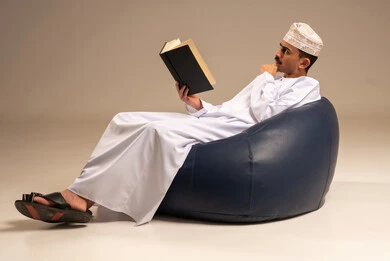 Reading stories and novels, a portrait of an Arab Gulf Omani man wearing traditional attire sitting on a comfortable chair holding a book he is reading, practicing the hobby of reading, enjoying beneficial recreational activities in free time, the concept of culture and self-learning, gray background.