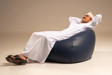 Relaxation and enjoying the calm atmosphere, the concept of elegance and attention to appearance, a portrait of a young Arab Gulf Omani man wearing a dishdasha and kumma sitting on a comfortable chair, with a gray background.