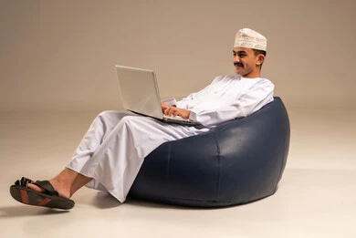 Remote work using a laptop, live streaming and making a video call via a computer, a portrait of an Arab Omani Gulf man wearing traditional attire sitting on a chair between badges and using a laptop, gray background.