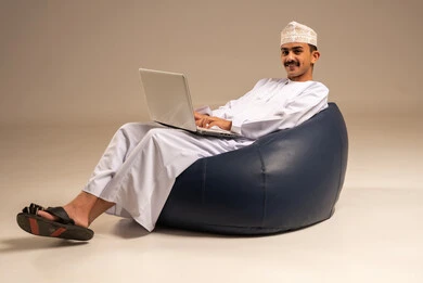 بث مباشر واجراء مكالمة فيديو عبر جهاز الكمبيوتر ، العمل عن بعد باستخدام اللابتوب ، بورتريه لرجل عربي عماني  خليجي يرتدي الزي التقليدي يجلس على كرسي بين بادج ويستخدم الحاسوب المحمول ، خلفية رمادية