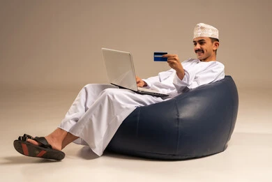 إدخال المعلومات البنكية على الحاسوب المحمول، توفير وسائل لتسهيل عملية الشراء والدفع عن بعد، بورتريه لرجل عربي  خليجي عماني يرتدي الثوب التقليدي يجلس على كرسي مريح يقوم باستخدام بطاقة الإئتمان في التسوق الالكتروني عبر الحاسب، خلفية رمادية