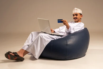 إدخال المعلومات البنكية على الحاسوب المحمول، توفير وسائل لتسهيل عملية الشراء والدفع عن بعد، بورتريه لرجل عربي  خليجي عماني يرتدي الثوب التقليدي يجلس على كرسي مريح يقوم باستخدام بطاقة الإئتمان في التسوق الالكتروني عبر الحاسب، خلفية رمادية