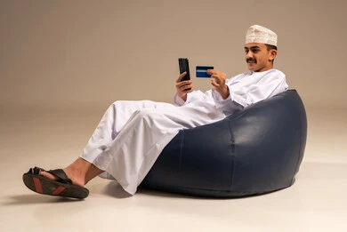A display of a blank black screen, entering banking information on a mobile phone, providing means to facilitate the process of remote purchasing and payment, a portrait of an Arab Omani Gulf man wearing traditional attire sitting on a comfortable chair using a credit card for online shopping via mobile, gray background.