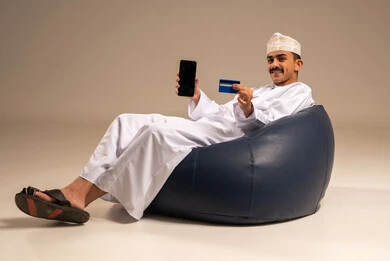 A display of a blank black screen, entering banking information on a mobile phone, providing means to facilitate the process of remote purchasing and payment, a portrait of an Arab Omani Gulf man wearing traditional attire sitting on a comfortable chair using a credit card for online shopping via mobile, gray background.