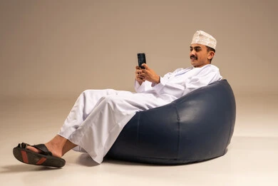 The use of modern devices and technologies, a portrait of an Arab Gulf Omani man wearing a dishdasha and kumma sitting on a chair in a comfortable lounge holding a mobile phone in his hand with gestures of joy, communicating with family and friends, browsing social media, gray background.