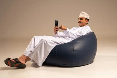 The use of modern devices and technologies, a portrait of an Arab Gulf Omani man wearing a dishdasha and kumma sitting on a chair in a comfortable lounge holding a mobile phone in his hand with gestures of joy, communicating with family and friends, browsing social media, gray background.