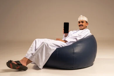 عرض شاشة سوداء فارغة عبر الهاتف، استخدام الأجهزة والتقنيات الحديثة، بورتريه لرجل عربي خليجي عماني يرتدي الدشداشة والكمة يجلس على كرسي بين بادج مريح يمسك الجوال بيده بإيماءات السرور، التواصل مع الاهل والاصدقاء، تصفح مواقع التواصل الاجتماعي، خلفية رمادية