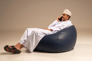 Making a phone call, using modern devices and technologies, a portrait of an Arab Gulf Omani man wearing a dishdasha and kumma sitting on a chair in a comfortable lounge holding a mobile phone in his hand with gestures of joy, communicating with family and friends, browsing social media, gray background.