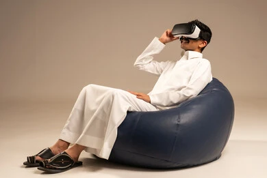 قضاء أوقات ممتعة ومسلية بالألعاب الافتراضية،الاستمتاع بمشاهدة أفلام الواقع الافتراضي،  بورتريه من الخلف لرجل عربي خليجي سعودي يرتدي الثوب التقليدي يجلس على كرسي مريح ويرتدي نظارة الواقع الافتراضي ثلاثية الابعاد، خلفية رمادية