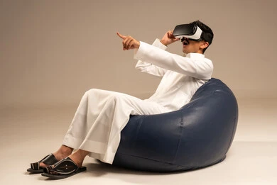 Interaction with virtual reality, a portrait of a young Arab Gulf Saudi man wearing traditional Saudi attire sitting on a chair among Bugs wearing 3D virtual reality glasses, enjoyment and entertainment in watching movies and simulating the virtual world, gray background.