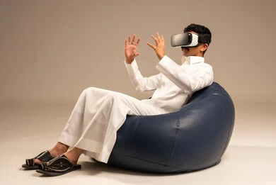 الاستمتاع بمشاهدة أفلام الواقع الافتراضي، قضاء أوقات ممتعة ومسلية بالألعاب الافتراضية، بورتريه من الخلف لرجل عربي خليجي سعودي يرتدي الثوب التقليدي يجلس على كرسي مريح ويرتدي نظارة الواقع الافتراضي ثلاثية الابعاد، خلفية رمادية