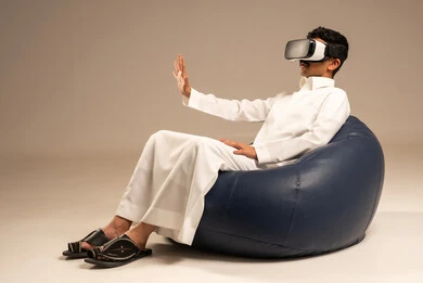 الاستمتاع بمشاهدة أفلام الواقع الافتراضي، قضاء أوقات ممتعة ومسلية بالألعاب الافتراضية، بورتريه من الخلف لرجل عربي خليجي سعودي يرتدي الثوب التقليدي يجلس على كرسي مريح ويرتدي نظارة الواقع الافتراضي ثلاثية الابعاد، خلفية رمادية
