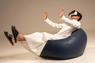 قضاء أوقات ممتعة ومسلية بالألعاب الافتراضية، الاستمتاع بمشاهدة أفلام الواقع الافتراضي، بورتريه من الخلف لرجل عربي خليجي سعودي يرتدي الثوب التقليدي يجلس على كرسي مريح ويرتدي نظارة الواقع الافتراضي ثلاثية الابعاد، خلفية رمادية