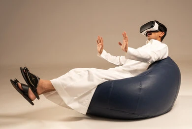 Spending enjoyable and entertaining times with virtual games, enjoying watching virtual reality movies, a portrait from behind of an Arab Gulf Saudi man wearing traditional attire sitting on a comfortable chair and wearing 3D virtual reality glasses, gray background.
