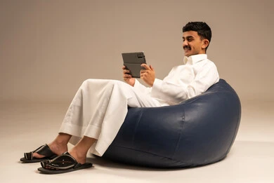 Communicating with family and friends via phone, using modern devices and technologies, a portrait of a Saudi Arabian Gulf man wearing traditional attire sitting on a chair in a comfortable lounge holding a tablet in his hand with gestures of joy, browsing social media, gray background.