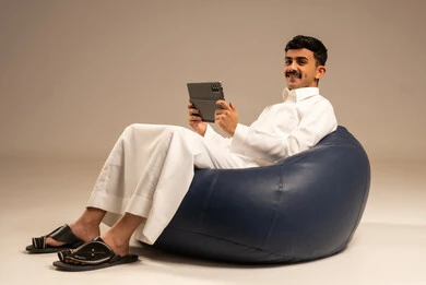 Communicating with family and friends via phone, using modern devices and technologies, a portrait of a Saudi Arabian Gulf man wearing traditional attire sitting on a chair in a comfortable lounge holding a tablet in his hand with gestures of joy, browsing social media, gray background.