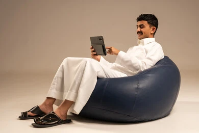 التواصل مع الاهل والاصدقاء عبر الهاتف، استخدام الأجهزة والتقنيات الحديثة، بورتريه لرجل عربي خليجي سعودي يرتدي الثوب التقليدي يجلس على كرسي بين بادج مريح يمسك التابلت بيده بإيماءات السرور، تصفح مواقع التواصل الاجتماعي، خلفية رمادية