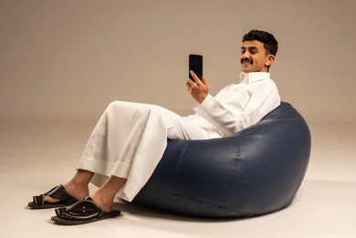 The use of modern devices and technologies, a portrait of a Gulf Arab Saudi man wearing a ghutrah and traditional thobe sitting on a chair in a comfortable lounge, holding a mobile phone in his hand with gestures of joy, communicating with family and friends, browsing social media, gray background.