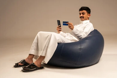 إدخال المعلومات البنكية على الهاتف المحمول، توفير وسائل لتسهيل عملية الشراء والدفع عن بعد، بورتريه لرجل عربي سعودي خليجي يرتدي الثوب التقليدي يجلس على كرسي مريح يقوم باستخدام بطاقة الإئتمان في التسوق الالكتروني عبر الجوال، خلفية رمادية