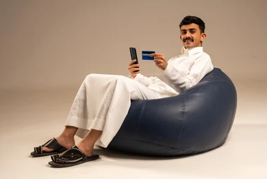 إدخال المعلومات البنكية على الهاتف المحمول، توفير وسائل لتسهيل عملية الشراء والدفع عن بعد، بورتريه لرجل عربي سعودي خليجي يرتدي الثوب التقليدي يجلس على كرسي مريح يقوم باستخدام بطاقة الإئتمان في التسوق الالكتروني عبر الجوال، خلفية رمادية