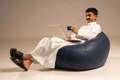 إدخال المعلومات البنكية على الحاسوب المحمول، توفير وسائل لتسهيل عملية الشراء والدفع عن بعد، بورتريه لرجل عربي سعودي خليجي يرتدي الثوب التقليدي يجلس على كرسي مريح يقوم باستخدام بطاقة الإئتمان في التسوق الالكتروني عبر الحاسب، خلفية رمادية