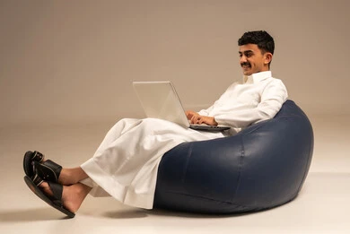 Live broadcast and making a video call via the computer, working remotely using a laptop, a portrait of an Arab Saudi Gulf man wearing traditional attire sitting on a chair between badges and using a laptop, gray background.