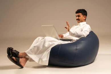 بث مباشر واجراء مكالمة فيديو عبر جهاز الكمبيوتر ، العمل عن بعد باستخدام اللابتوب ، بورتريه لرجل عربي سعودي خليجي يرتدي الزي التقليدي يجلس على كرسي بين بادج ويستخدم الحاسوب المحمول ، خلفية رمادية