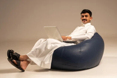 بث مباشر واجراء مكالمة فيديو عبر جهاز الكمبيوتر ، العمل عن بعد باستخدام اللابتوب ، بورتريه لرجل عربي سعودي خليجي يرتدي الزي التقليدي يجلس على كرسي بين بادج ويستخدم الحاسوب المحمول ، خلفية رمادية