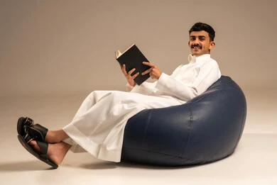 The concept of culture and self-learning, reading with contemplation and focus, spending free time learning, a portrait of an Arab Saudi Gulf man wearing traditional attire sitting on a comfortable chair holding a scientific educational book, gray background.