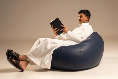 القراءة بتمعن وتركيز، قضاء وقت الفراغ بالتعلم، مفهوم الثقافة والتعلم الذاتي، بورتريه لرجل عربي سعودي خليجي مبتسم يرتدي الثوب التقليدي يجلس على كرسي مريح يحمل بيده كتاب علمي تثقيفي، خلفية رمادية