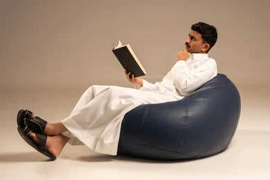 القراءة بتمعن وتركيز، قضاء وقت الفراغ بالتعلم، مفهوم الثقافة والتعلم الذاتي، بورتريه لرجل عربي سعودي خليجي مبتسم يرتدي الثوب التقليدي يجلس على كرسي مريح يحمل بيده كتاب علمي تثقيفي، خلفية رمادية