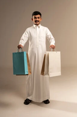 متعة التسوق في المملكة, التبضع وأخذ الحاجات اللازمة, المشتريات التجارية والهدايا, مواسم التخفيضات في السعودية, الاستفادة من الحسومات والتصفيات, بورتريه لشاب عربي خليجي سعودي يرتدي الثوب التقليدي يمسك بيده أكياس ورقية ملونة,  الجسم بالطول الكامل, خلفية رمادية