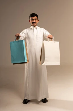 Shopping and taking necessary items, commercial purchases and gifts, discount seasons in Saudi Arabia, benefiting from discounts and clearance sales, a portrait of a young Arab Gulf Saudi man wearing traditional attire holding colorful paper bags, the joy of shopping in the Kingdom, full-body, gray background.