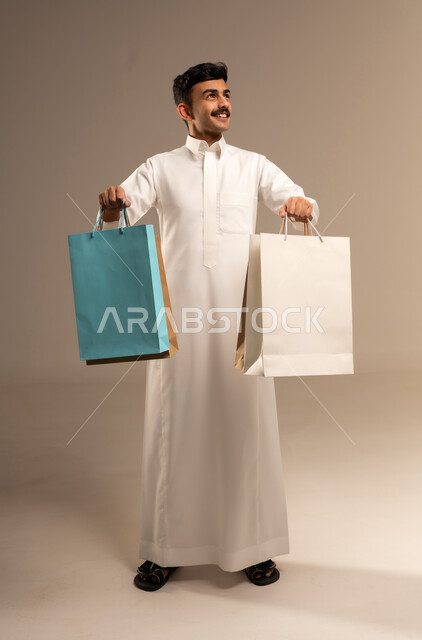 The joy of shopping in the Kingdom, buying and taking necessary items, commercial purchases and gifts, discount seasons in Saudi Arabia, benefiting from discounts and clearance sales, a portrait of a young Arab Gulf Saudi man wearing traditional attire holding colorful paper bags in his hand, full-body shot, gray background.