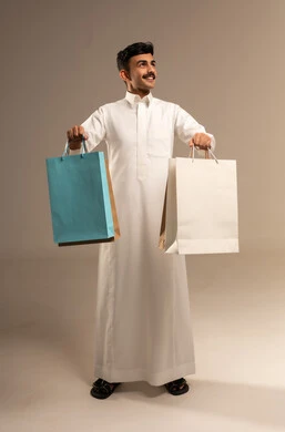 The joy of shopping in the Kingdom, buying and taking necessary items, commercial purchases and gifts, discount seasons in Saudi Arabia, benefiting from discounts and clearance sales, a portrait of a young Arab Gulf Saudi man wearing traditional attire holding colorful paper bags in his hand, full-body shot, gray background.
