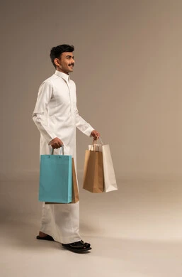 Shopping and taking necessary items, commercial purchases and gifts, discount seasons in Saudi Arabia, benefiting from discounts and clearance sales, a portrait of a young Arab Gulf Saudi man wearing traditional attire holding colorful paper bags, the joy of shopping in the Kingdom, full-body, gray background.