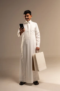 Completing the ordering process using a mobile phone, the concept of remote shopping, online electronic purchases, a portrait of a young Arab Gulf Saudi man wearing traditional attire holding colorful paper bags, shopping and taking advantage of offers and discounts, full-body, gray background.
